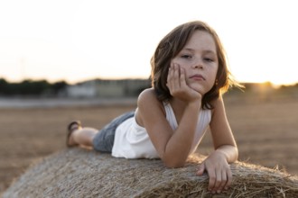 Child in casual attire lies on a hay bale during sunset, reflecting a calm and serene mood. The