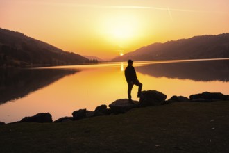 A person standing on rocks with a view of a calm lake at sunset, Großer Alpsee, Immenstadt im