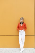 A fashionable woman leans against a striking orange wall in Warsaw, wearing bright attire with