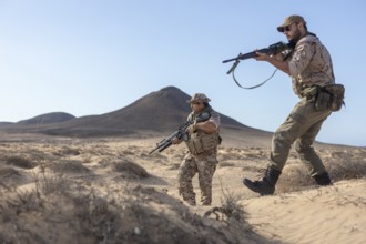 Two soldiers in camouflage gear patrol a sandy desert holding rifles. They observe their