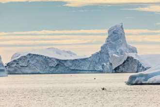 A large iceberg protrudes from the sea, a kayaker small below. The sky is cloudy, icebergs in the