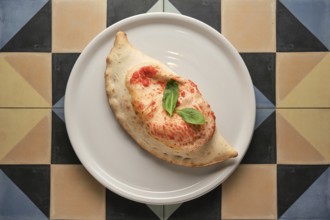 Freshly baked calzone garnished with basil leaves, served on a white plate over a vibrant geometric