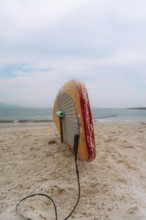 A surfboard lies on the sandy shore of a Peruvian beach, tethered by its leash, under a cloudy sky.