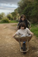 Two children enjoying a playful moment with a wheelbarrow on a rural path. The older child pushes