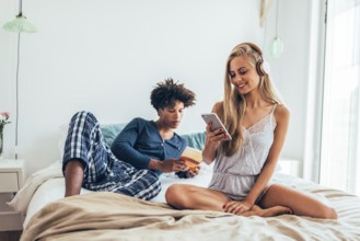 A couple enjoys a relaxed morning on a cozy bed, one listens to music on headphones, the other