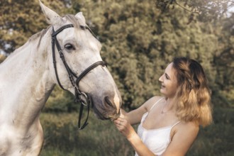 A young woman gently interacts with a white horse in a tranquil outdoor countryside environment.