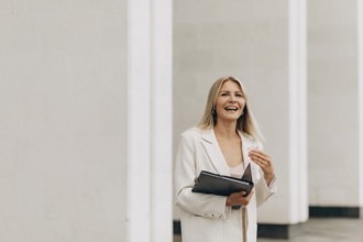 A businesswoman in a white suit smiles joyfully while holding a folder. She stands in front of