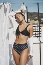 A young woman in a black polka-dot bikini and white shirt enjoys the summer sun on a rooftop The