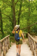 A woman wearing a straw hat and a green backpack is walking across a bridge. The bridge is