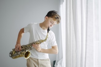 A young boy plays the saxophone in natural light by a large window. The casual outfit and serene