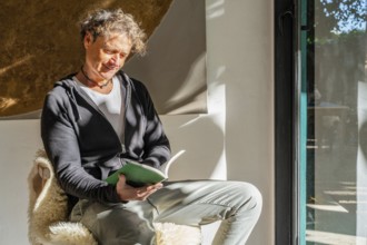 A man enjoys a quiet moment reading a book at home, sitting comfortably by a window, illuminated by