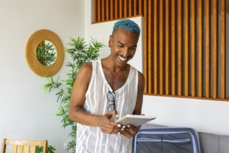 Hispanic LGBTQIA+ man with blue hair happily prepares for holidays using a tablet, surrounded by