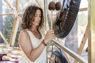A female sound therapist plays a large gong during a gong bath session with eyes closed. The serene