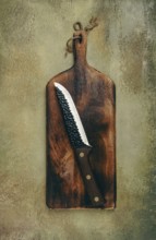 A rustic wooden cutting board displays a sharp knife resting diagonally. The textured background