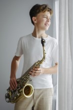 A young saxophonist in a white shirt looks out the window, standing in soft sunlight. The relaxed