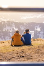 Couple sitting on a mountain peak with a view of the sunny winter valley, Hochgrat Mountains,