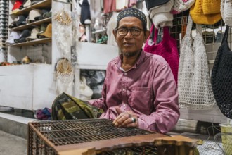 A balinese vendor in a traditional market, wearing glasses. Surrounded by various handmade items