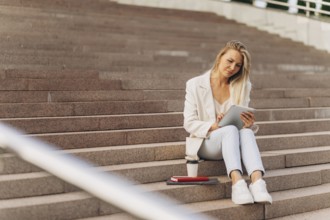 A woman in a white blazer and jeans sits on outdoor steps, using a tablet. Beside her are a