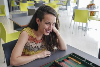 A woman sits at a table in a modern cafe, smiling and engaged, while playing a game of backgammon