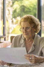 Elderly woman with glasses reading documents at home, sitting at a table with natural light coming
