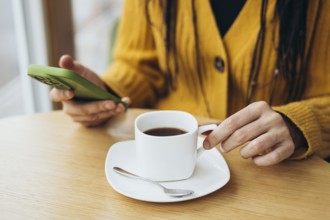 A person in a yellow sweater holds a smartphone while enjoying coffee at a wooden table A white