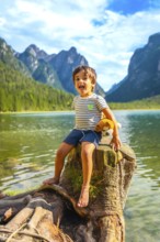 Carefree child sticking out tongue, sitting on tree stump by lake dobbiaco with plush toy, enjoying