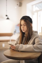 A teenage girl wearing a hoodie and cap sits at a small table, engrossed in her smartphone. The