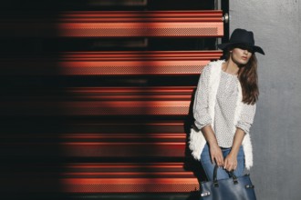 A fashionable woman in a wide-brimmed hat and stylish clothing poses with a handbag against a red