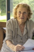 Portrait of a senior woman sitting at a table on a sunny veranda, holding and reading a book,