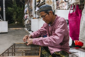 A balinese artisan focuses intently while crafting at a market stall. Surrounded by handmade