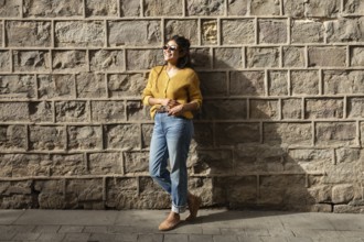 A Latin woman in casual attire, wearing sunglasses, smiles while leaning against a sunlit brick