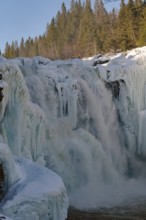 A frozen waterfall with forest in the background and snow on the rocks, water roaring over