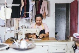A asian seamstress works on a sewing machine in a cozy workshop. Surrounded by hanging clothes and