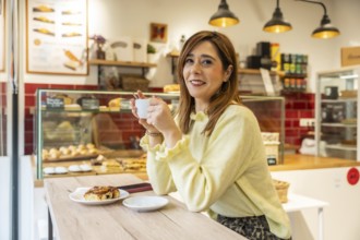 Young woman in a light yellow sweater smiles at a sunny cafe counter, holding espresso and enjoying