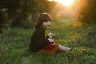 A young child sits on lush grass with orange trees, playing a red ukulele in a sunlit meadow. The