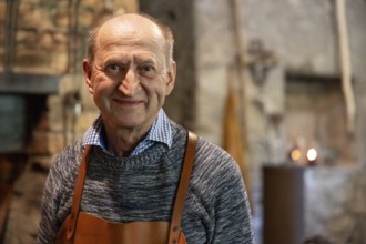 A skilled blacksmith wearing a leather apron smiles warmly in his rustic workshop. The background