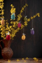 Branches of forsythia (Forsythia) in a rustic jug hung with Easter eggs against a dark background