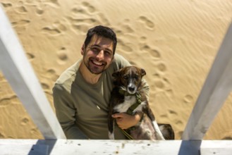 A cheerful man holding his dog on a sunny beach in Greece. The sand is marked with footprints.