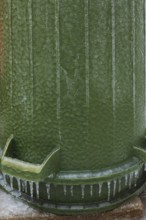 Upside down green PVC and rubber thrash can covered with thick sheet of ice and hanging icicles