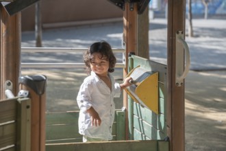 A cheerful toddler in a white shirt explores a playground on a sunny day, engaging with playful