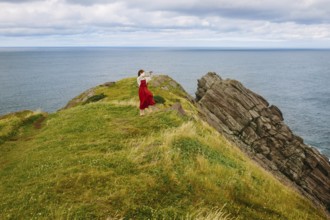 A woman clad in a flowing red dress stands on the rugged cliffs of Middle Cove overlooking the