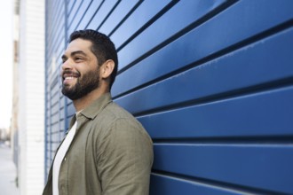 A bearded man with a friendly smile leans against a blue wall on an urban street. He has short hair