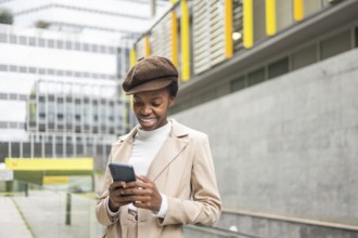 An urban black woman smiles while checking her phone in the city She wears a beige coat and cap,