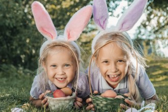 Two smiling children wearing bunny ears enjoy an Easter egg hunt in a sunny garden, holding baskets