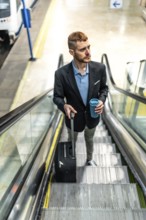 Businessman going up escalator in subway station, holding reusable coffee cup and pulling trolley