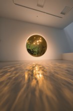 A round mirror on the wall casts warm light reflections onto the floor in an art space, Schauwerk