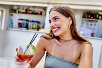 A woman smiles while holding a colorful cocktail adorned with mint at a lively beach bar. The