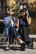 A multiethnic lesbian couple sits on a bench outdoors, wearing headphones and sharing a joyful