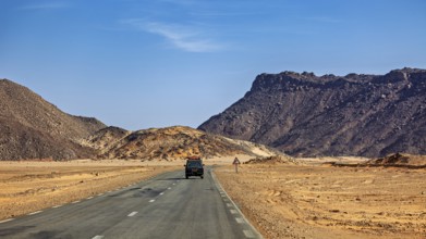 An off-road vehicle drives on a tarmac road through a mountainous desert landscape, With an