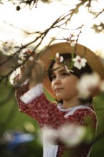 A young girl in a red dress and straw hat looks at an almond tree in bloom on a sunny day. The